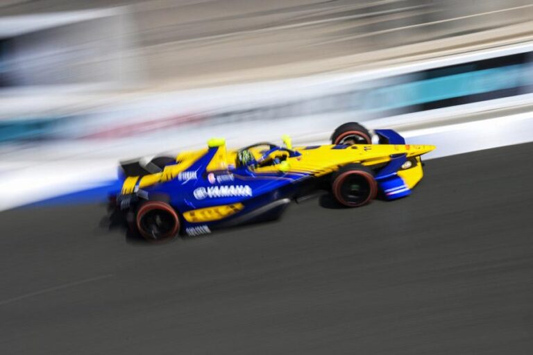 Fotografia de ação com desfoque de movimento de um carro Gen3 da Fórmula E da Lola Yamaha Abt (amarelo e azul, carro nº 11, Lucas Di Grassi) correndo em alta velocidade no circuito 'old school' de Jarama, Espanha, para o ePrix de Madri. A pista tem asfalto áspero e barreiras tradicionais, com placas indicativas visíveis.